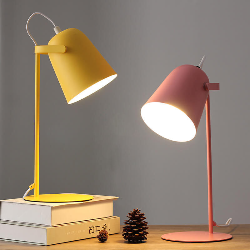 Zwei leuchtende Tischlampen im Vergleich, eine gelbe auf Büchern und eine rosafarbene auf dem Holztisch, dazwischen ein Tannenzapfen. 5