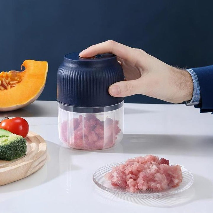 Eine Hand drückt den dunkelblauen Deckel des Mini-Zerkleinerers, im Behälter steckt zerkleinertes Fleisch, daneben ein Teller mit Hackfleisch und Gemüse. 1