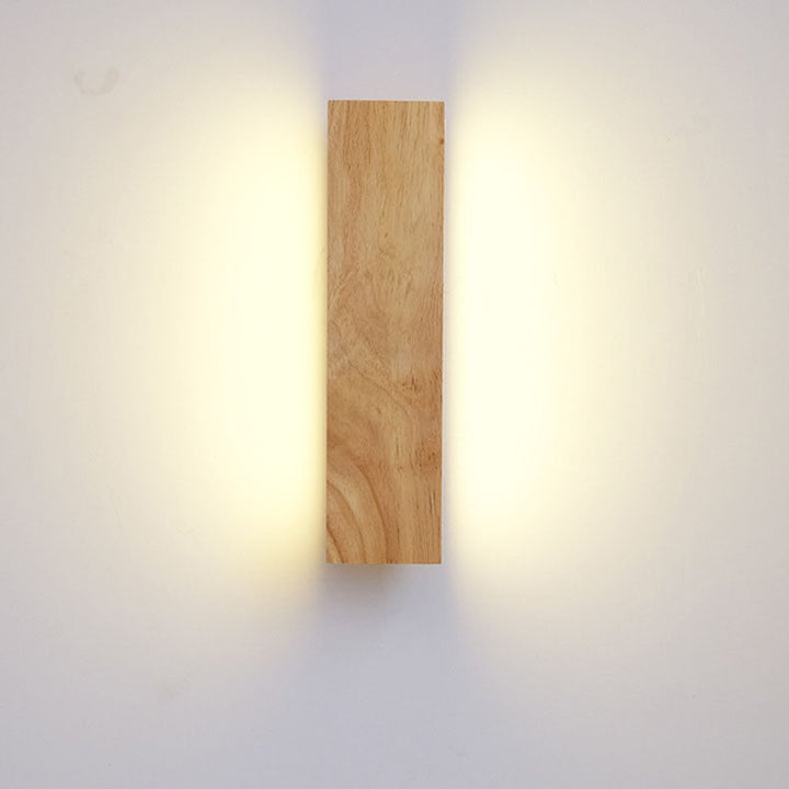 Frontansicht der senkrecht hängenden Holzleuchte, ein warmer Lichtschein fällt seitlich links und rechts auf die Wand. 3