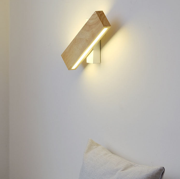Holz-Wandleuchte mit weißer Halterung, schräg gedreht und eingeschaltet, warmes Licht an heller Wand über einem Kissen. 1