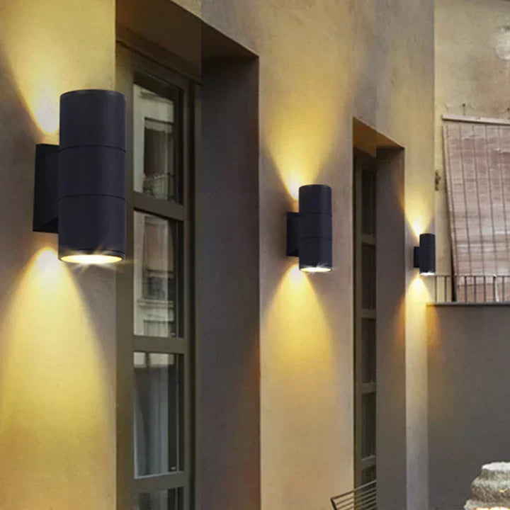 Drei schwarze Wandleuchten in unterschiedlichen Größen an einer Hausfassade neben Fenstern, die warmes Licht an die Wand strahlen. 3