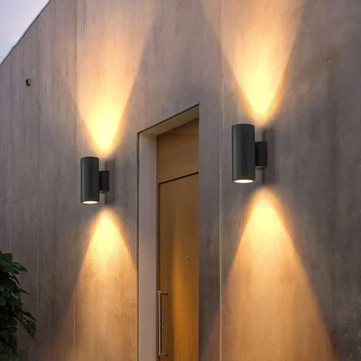 Zwei schwarze Wandleuchten links und rechts neben einer Holztür, die warme Lichtkegel nach oben und unten auf die Betonfassade werfen. 6