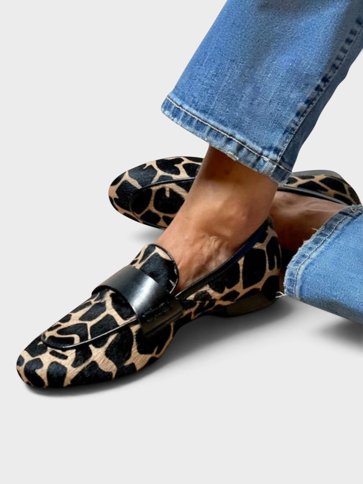 Flache Slip-on-Loafer mit schwarz-beigem Fellmuster und schwarzem Lederband, getragen zu Jeans, die Beine locker übereinandergeschlagen, Schrägansicht von oben. 1