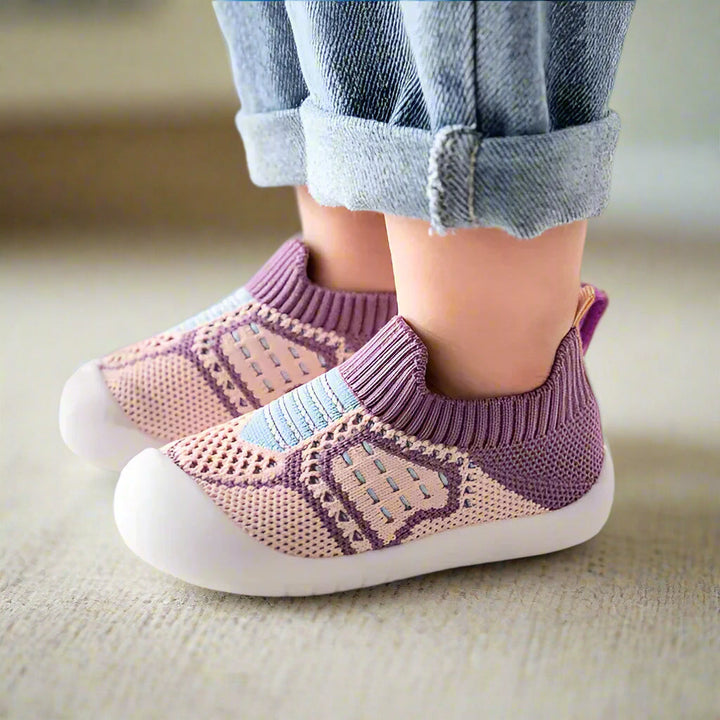 Kinderfüße in lila-rosafarbenen Strickschuhen mit hellblauen Streifen und weißer Gummisohle auf hellem Teppich. 10