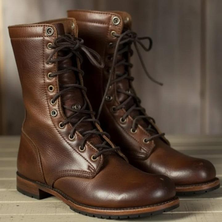 Paar hohe braune Lederstiefel mit dunklen Schnürsenkeln und Metallösen, schräg von vorn auf einem hellen Holzboden fotografiert. 1