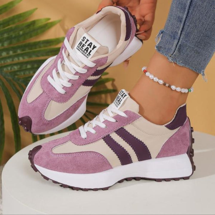 Rosa-beige Sneaker mit violetten Streifen, ein Schuh angehoben, auf weißem Sockel vor Palmenblatt 11