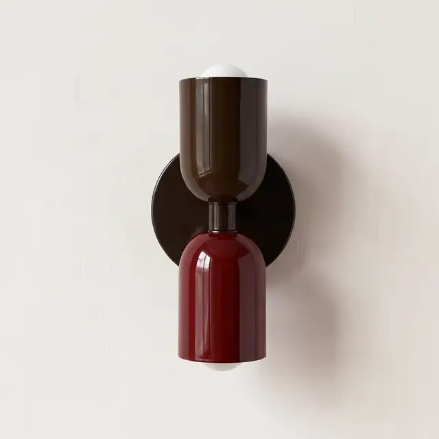 Wandlampe mit dunkelbraunem oberem Schirm, weinrotem unterem Schirm und dunkler runder Wandplatte vor heller Wand. 24