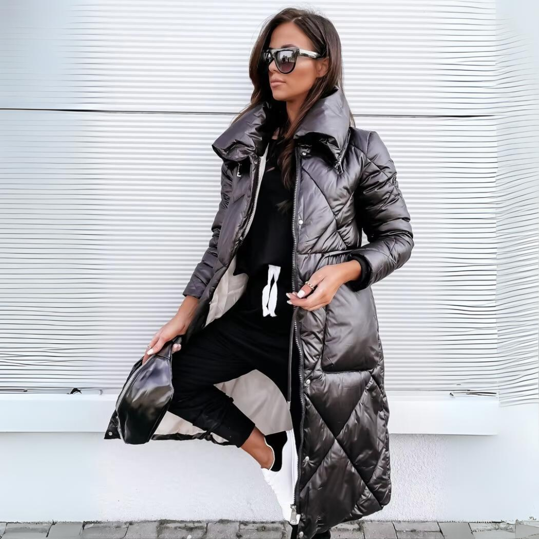 Frau mit Sonnenbrille trägt die lange, glänzend schwarze Steppjacke mit hohem Kragen offen über schwarzem Outfit und hält eine kleine Tasche. 1