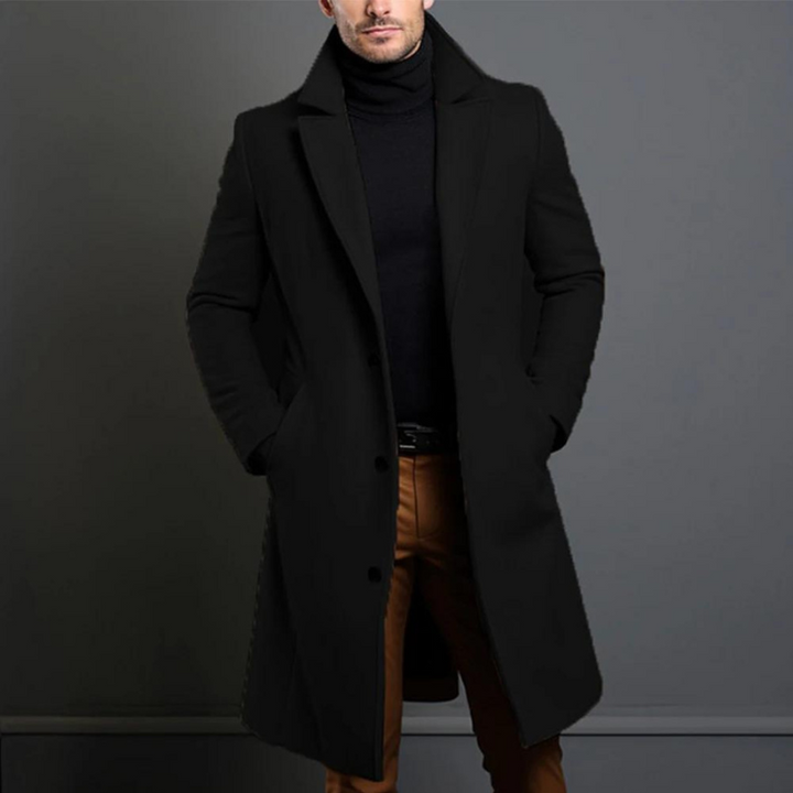 Schwarzer langer Mantel mit Reverskragen und Knopfleiste, getragen mit schwarzem Rollkragenpullover und braunen Hosen, Hände in den Taschen. 1