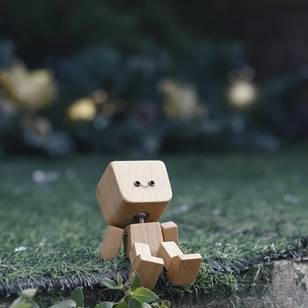 Holzfigur mit freundlichem Gesicht sitzt draußen auf Kunstrasen, im Hintergrund unscharfe goldene Lichter. 5