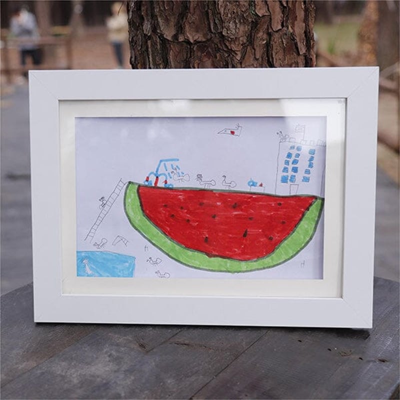 Weißer Bilderrahmen mit Kinderzeichnung einer riesigen Wassermelone, im Freien auf einem Holztisch vor einem Baumstamm aufgestellt. 21