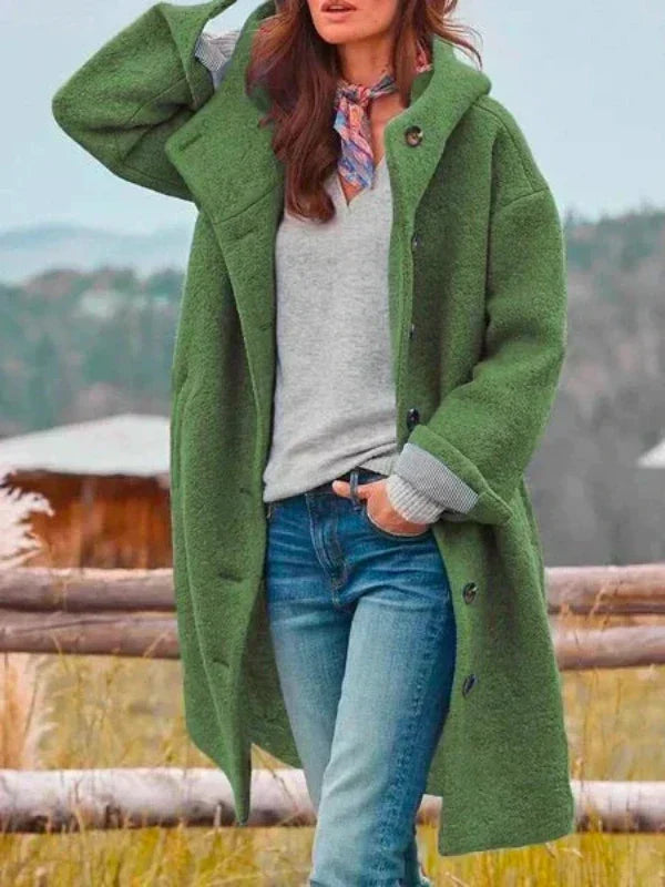 Langer Kapuzenmantel in Grün, offen getragen über grauem Pullover und Jeans, mit buntem Halstuch vor ländlicher Kulisse. 5