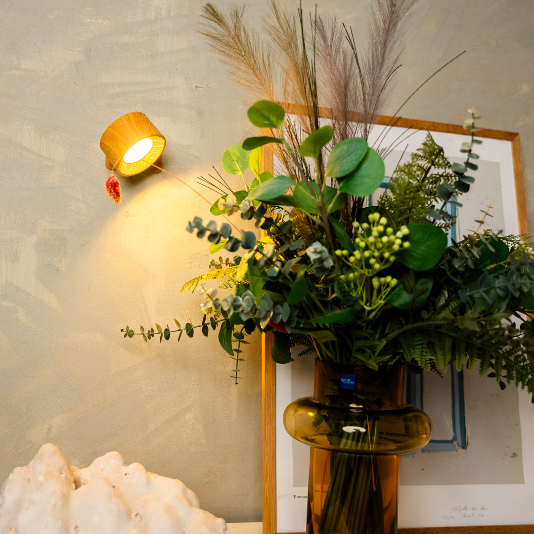 Wandlampe in Holzoptik leuchtet warm neben einem Strauß aus Grünzweigen und Pampasgras in einer Bernsteinvase. 6