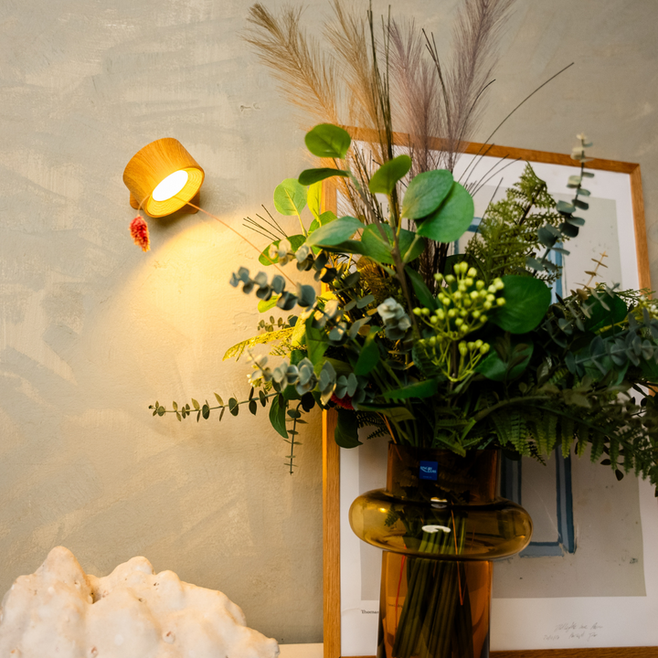 Wandlampe in Holzoptik leuchtet warm neben einem Strauß aus Grünzweigen und Pampasgras in einer Bernsteinvase. 6