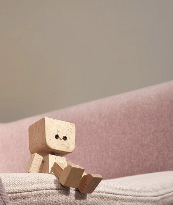 Kleine Holzfigur mit lächelndem Gesicht sitzt auf der Kante eines rosa Polstersofas vor einer grauen Wand. 4