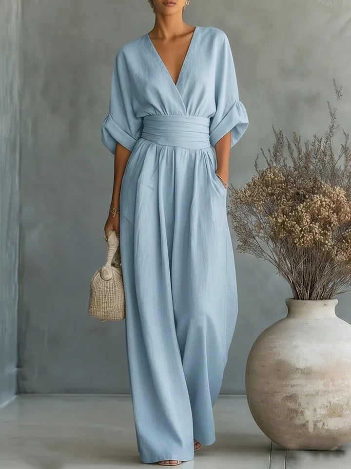 Frontansicht des Jumpsuits in Hellblau mit V-Ausschnitt, betonter Taille und weitem Hosenbein, dazu eine helle Strohtasche. 2