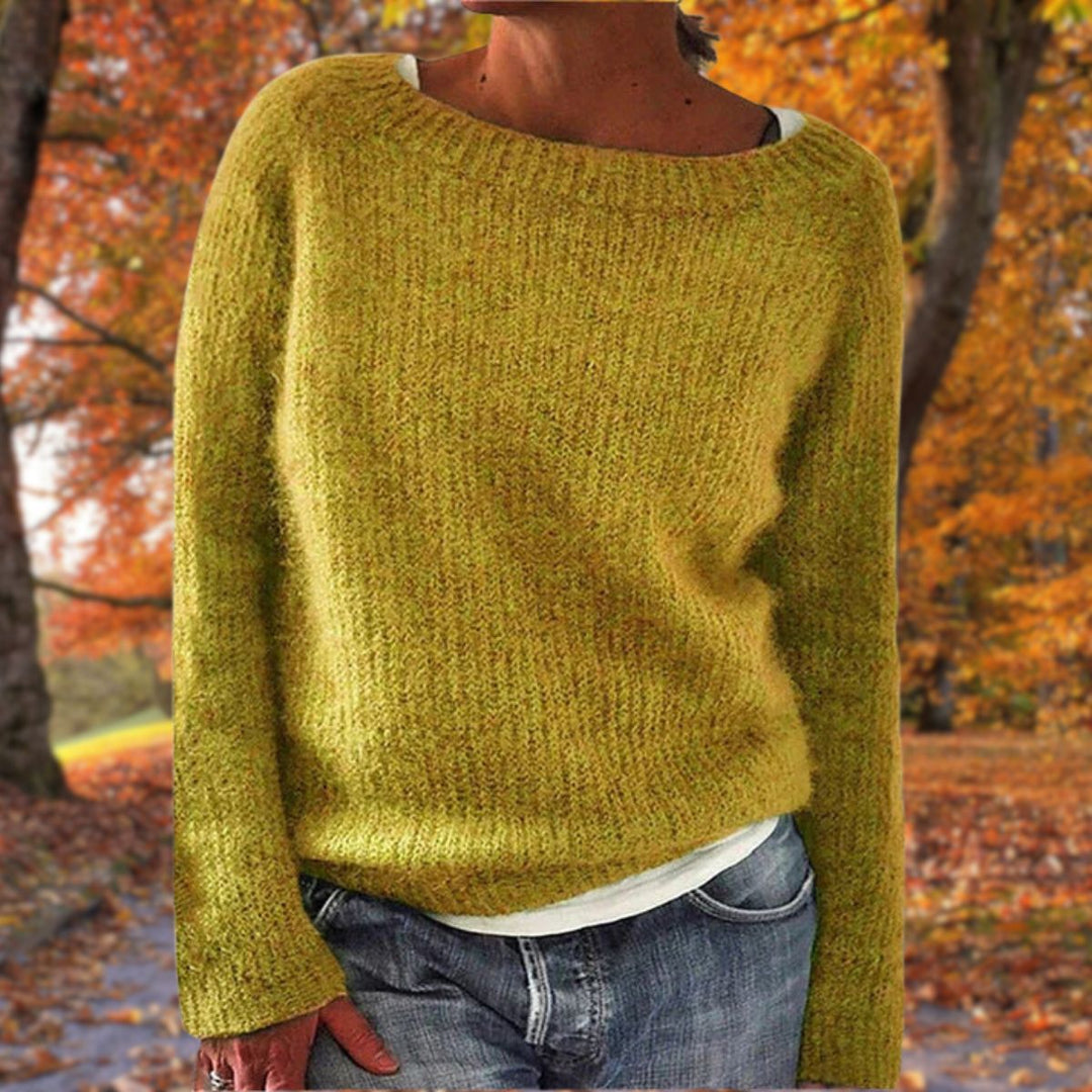 Senfgelber Strickpullover mit Rundhalsausschnitt, getragen über einem weißen Shirt und Jeans vor herbstlichem Laubwald. 1