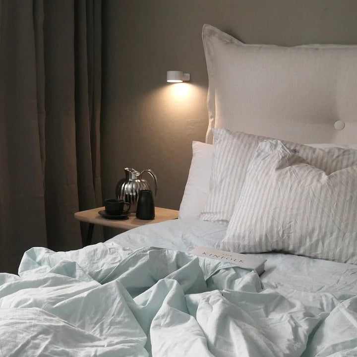 Kleine weiße Wandlampe über dem Nachttisch eines Bettes mit hellblauer Bettwäsche spendet gedämpftes Licht. 7