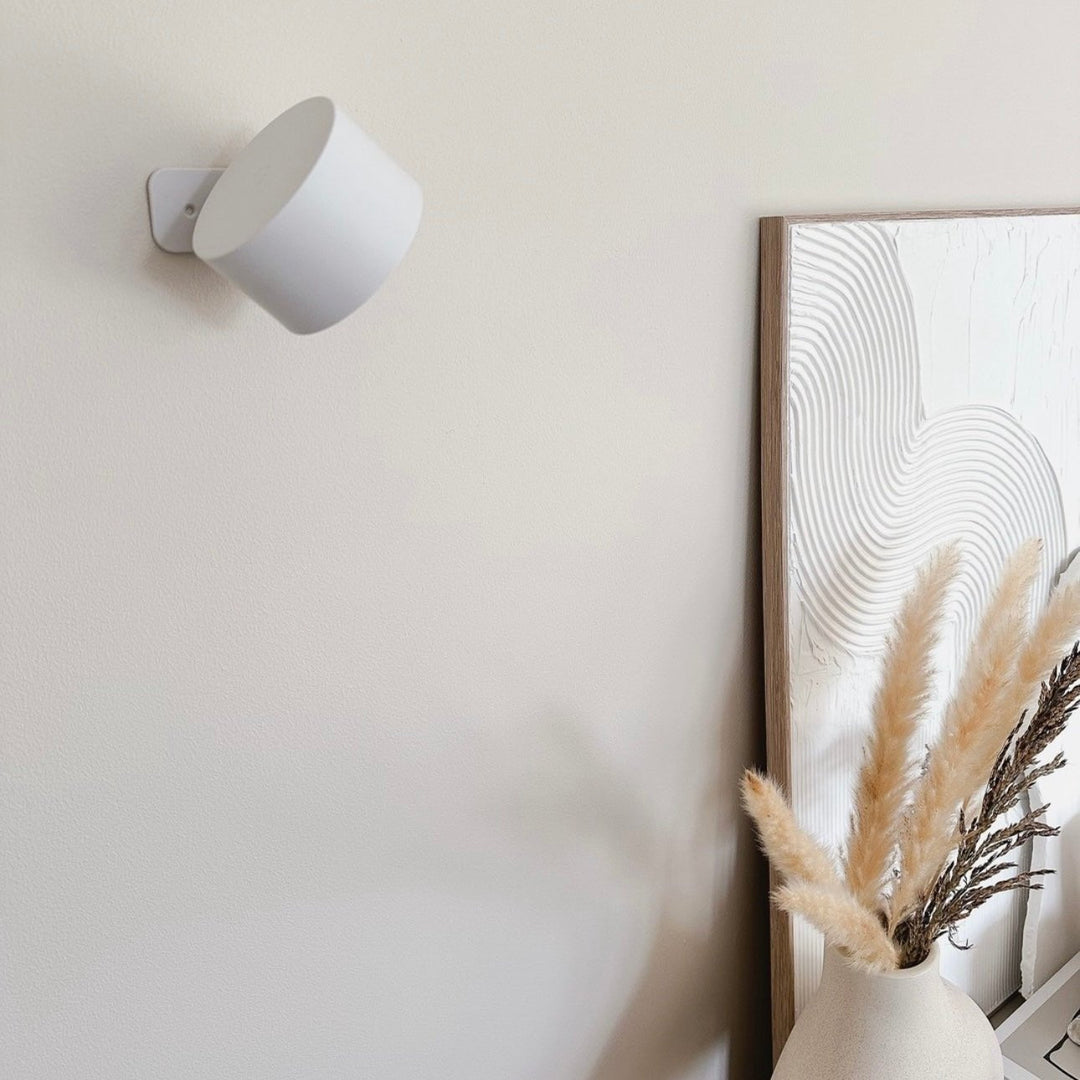 Ausgeschaltete weiße Wandlampe an einer beigen Wand neben einem strukturierten Bild und Pampasgras in einer Vase. 17