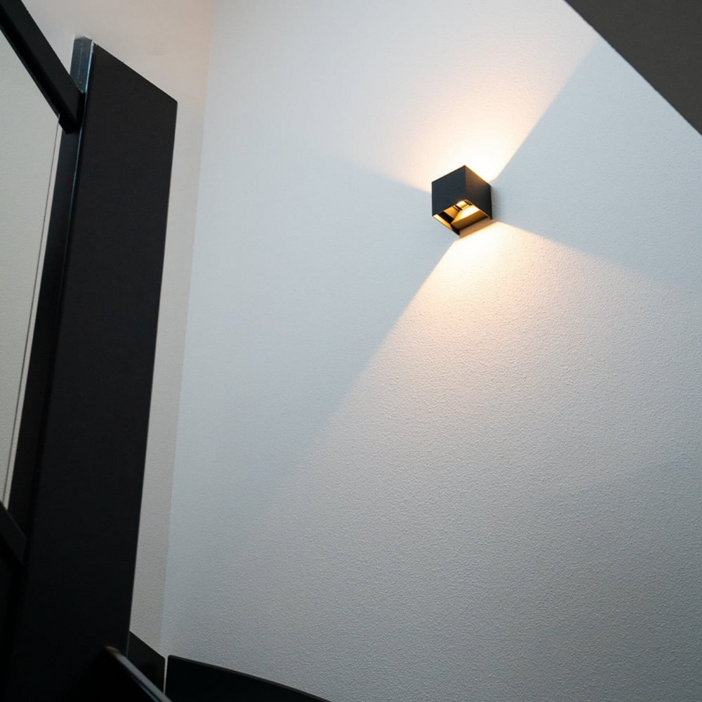Schwarze Würfelleuchte an einer hellen Treppenhauswand, von unten fotografiert, wirft warmes Licht in einem Kegel nach oben und unten. 2