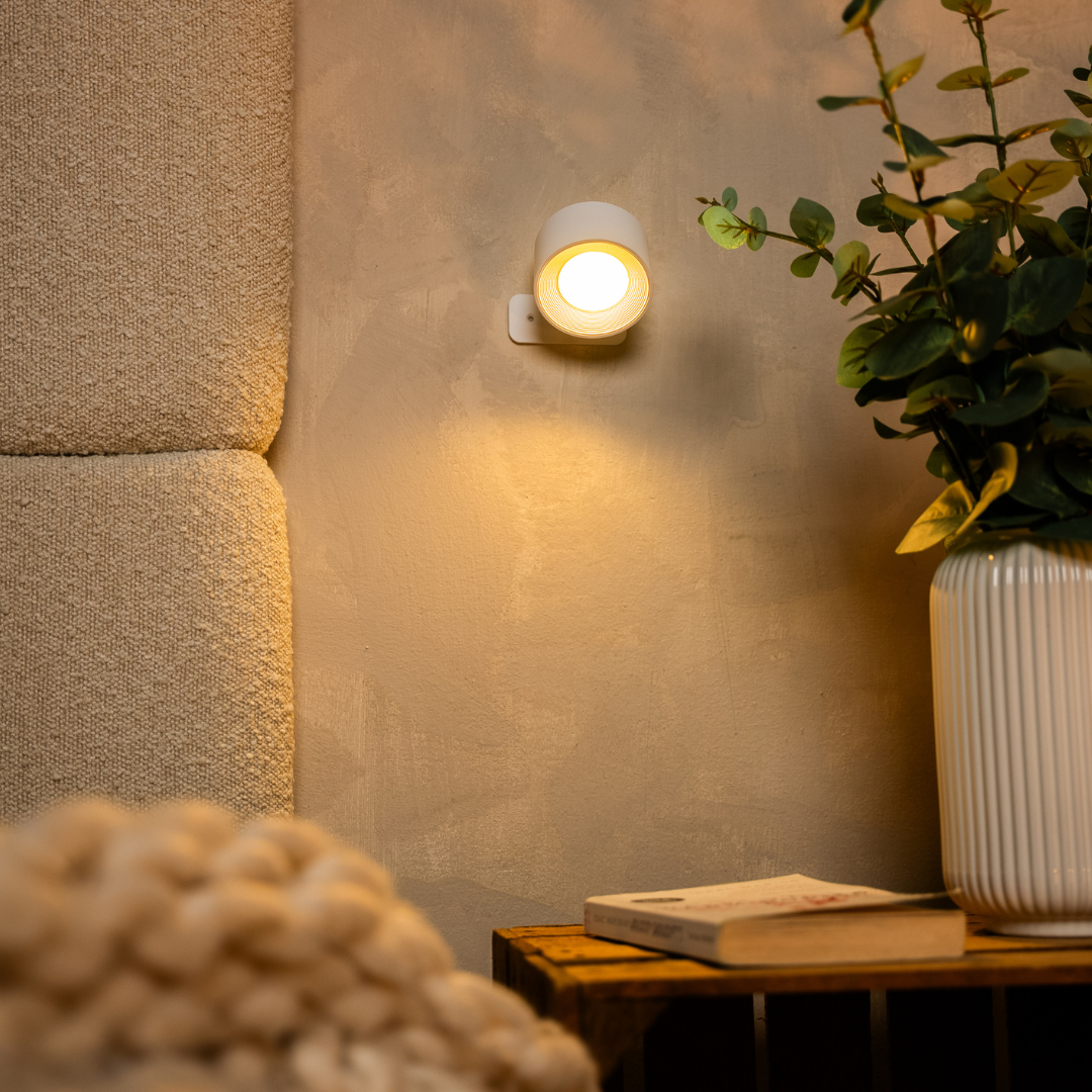 Weiße Wandlampe leuchtet warm an einer verputzten Wand neben einer Vase mit Eukalyptuszweigen und einem Buch auf einer Holzkiste. 12