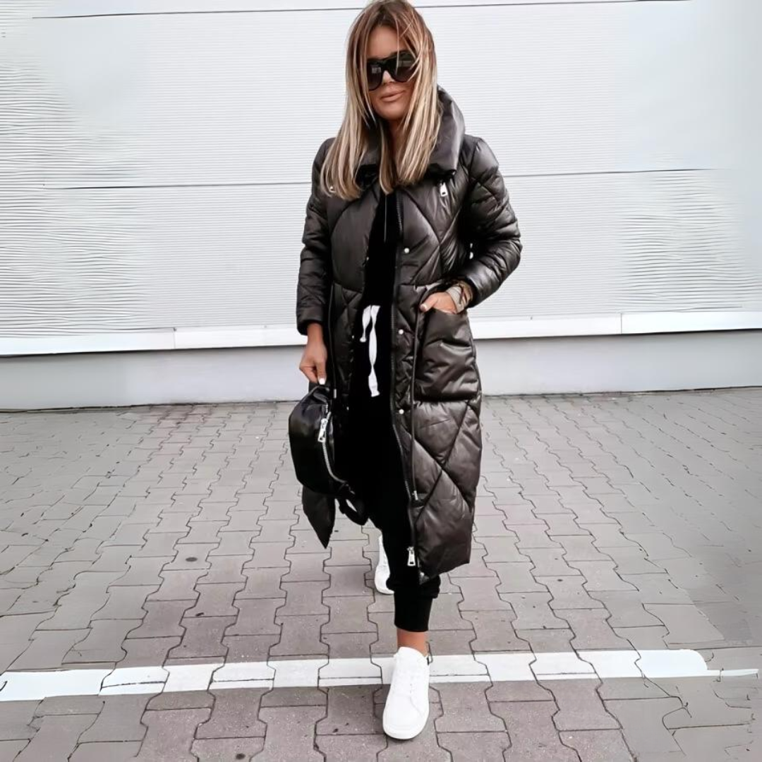 Frau mit Sonnenbrille steht auf Pflaster und trägt die lange schwarze Steppjacke geschlossen, eine Hand in der Seitentasche. 5