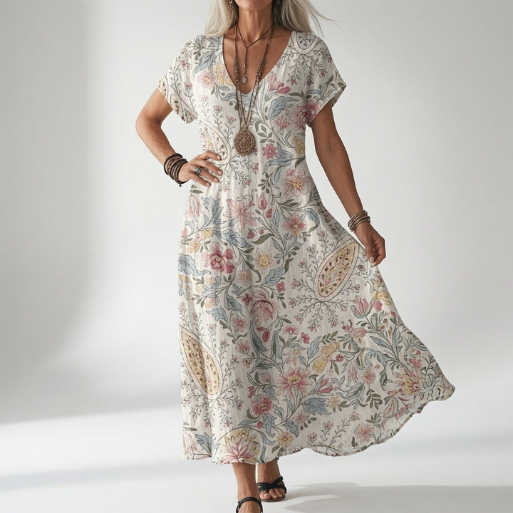 Frau trägt das Kleid in Cremeweiß mit zartem Blumen- und Paisleymuster in Rosa und Blau, kombiniert mit langer Holzkette und Sandalen. 1
