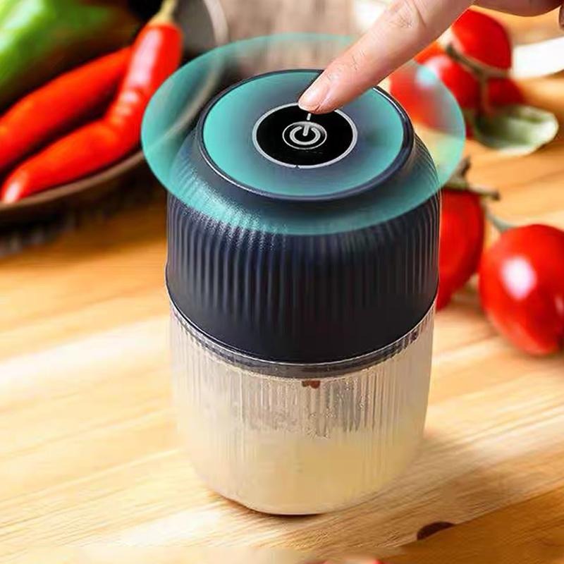 Nahaufnahme von oben: Ein Finger tippt auf die runde Einschalttaste am gerillten dunkelblauen Deckel, im Hintergrund Chilischoten und Tomaten. 2