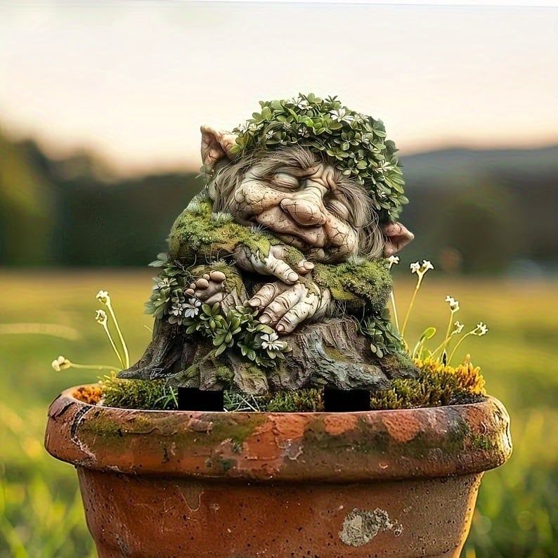 Schlafender Troll mit Blätterkranz in einem Terrakotta-Topf mit Moos und kleinen weißen Blüten, vor Wiese im Abendlicht. 17