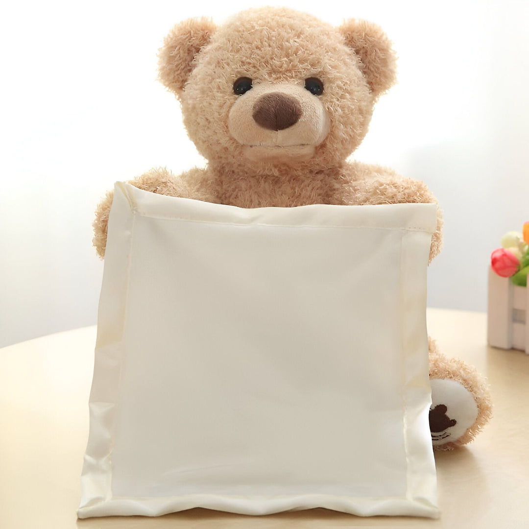 Frontalansicht des hellbraunen Plüschbären, der mit beiden Armen ein cremefarbenes Satintuch vor seinen Körper hält, auf einem Tisch. 2