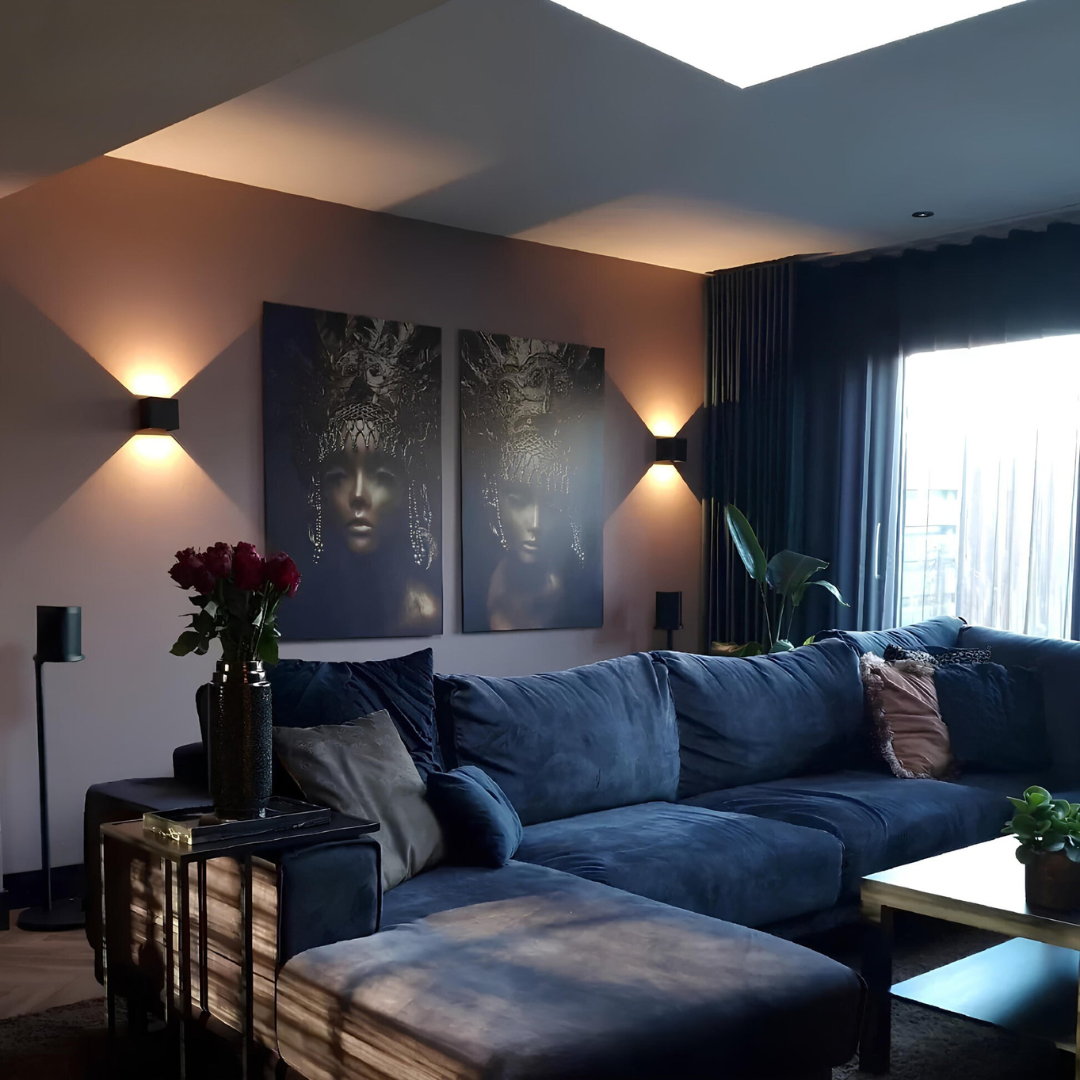 Zwei schwarze Wandleuchten über einem blauen Samtsofa im Wohnzimmer, zwischen ihnen zwei dunkle Gemälde, rechts ein Vorhang. 9