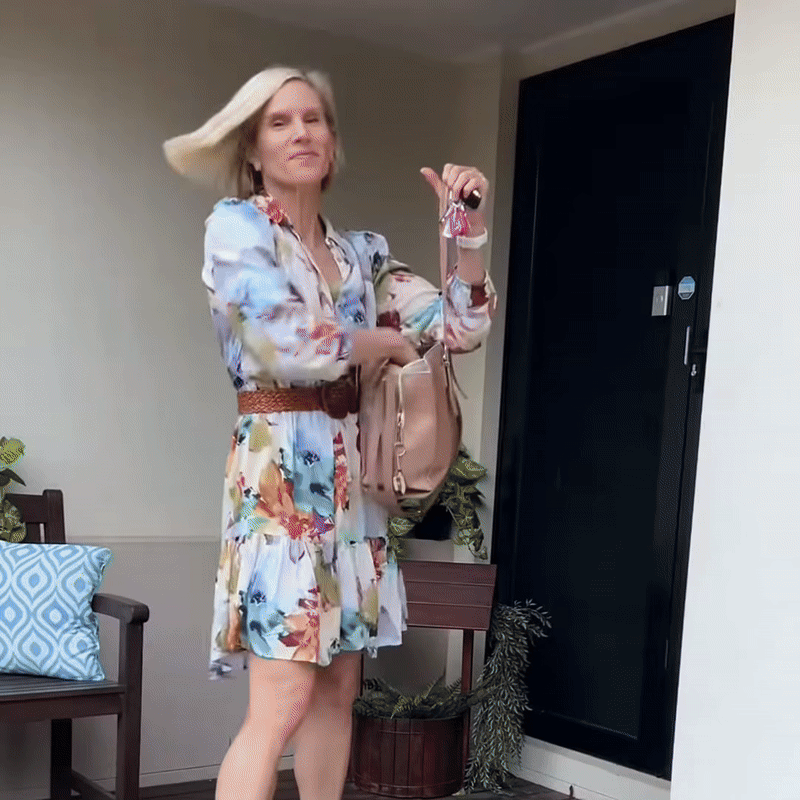 Blonde Frau in geblümtem Kleid mit Gürtel und Handtasche schwingt ihr glattes Haar vor einer dunklen Haustür. 8
