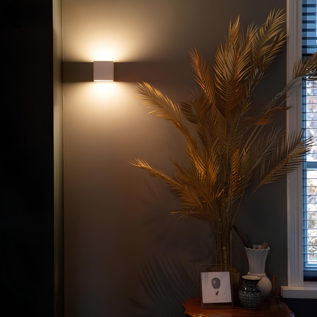 Weiße Würfelleuchte an einer dunklen Wand neben einer Vase mit goldenen Palmwedeln und einem Fenster, warmes Licht nach oben. 4