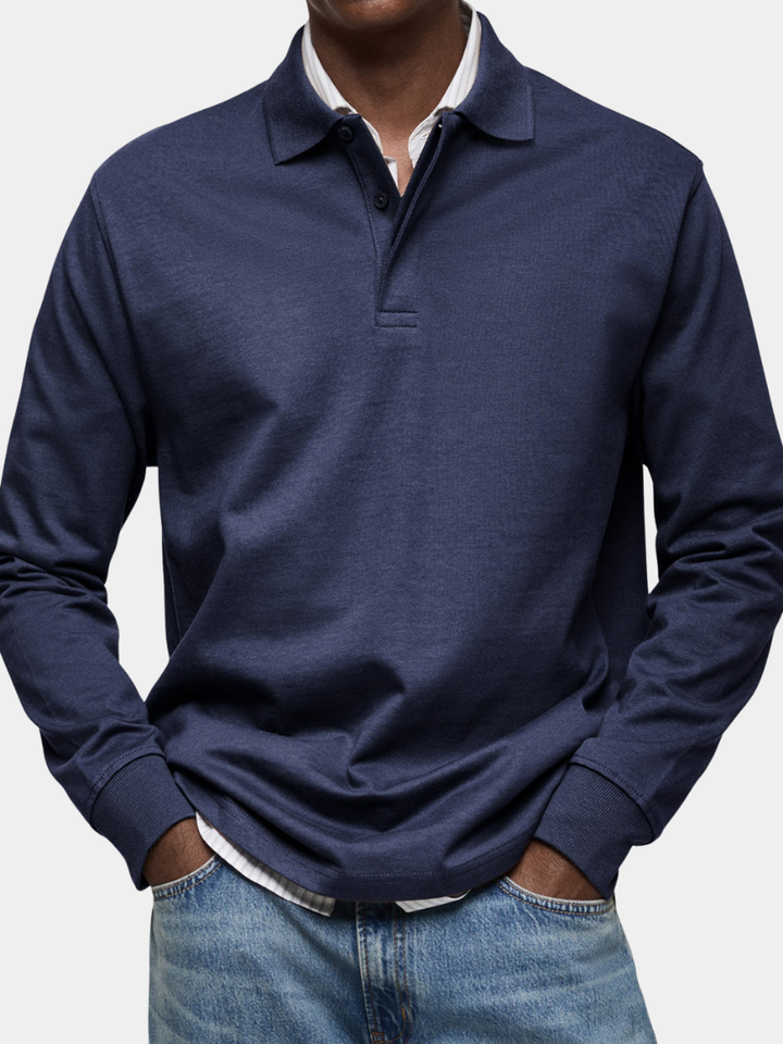 Nahaufnahme des marineblauen Langarm-Poloshirts am Model, kombiniert mit gestreiftem Hemd darunter und Jeans, Hände in den Taschen. 1