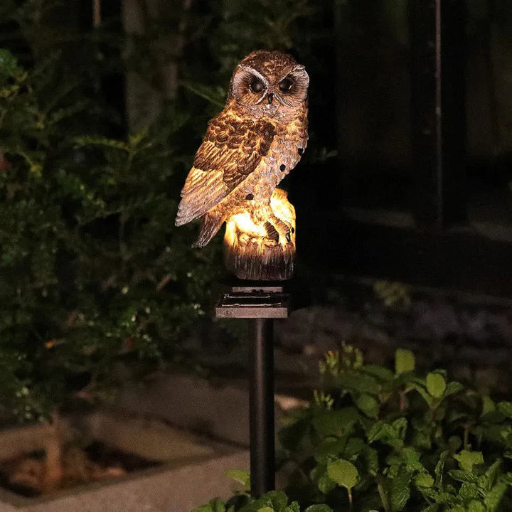 Braune Eule auf Baumstumpf mit Spieß, abends warm leuchtend im dunklen Garten zwischen Büschen und Blattpflanzen. 4