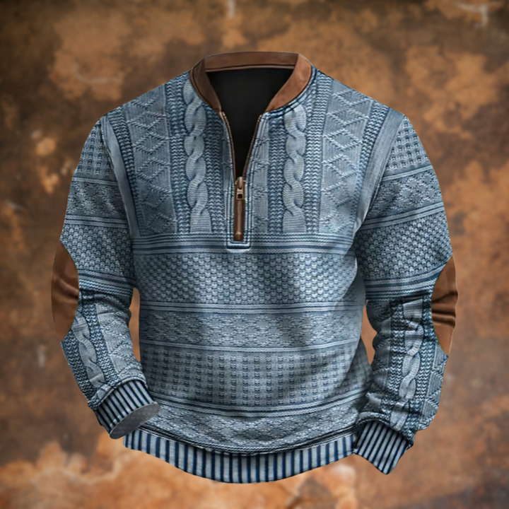 Frontansicht des Strickpullovers in Blaugrau mit Zopfmuster, braunem Stehkragen, Reißverschluss am Ausschnitt und braunen Ellbogenpatches vor rostigem Hintergru 1