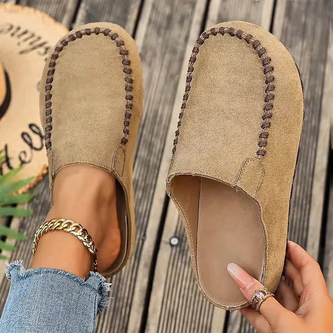 Beigefarbene Slipper mit brauner Ziernaht von oben auf Holzdielen, ein Schuh am Fuß, am anderen das Fußbett sichtbar. 2