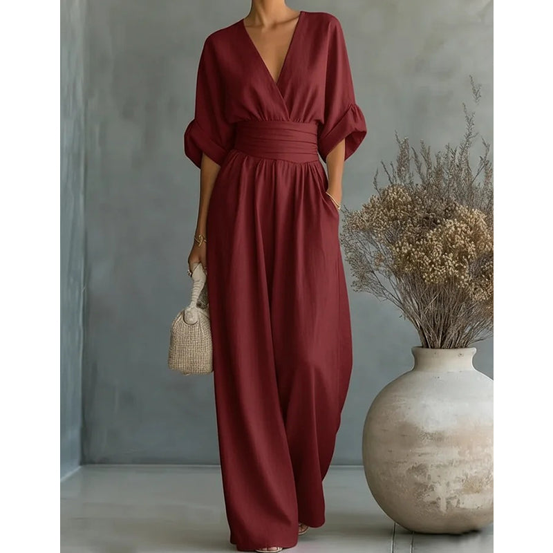 Weinroter Jumpsuit in der Frontansicht mit V-Ausschnitt, hohem Taillenband und weitem Bein, Hand in der Tasche, vor grauem Hintergrund. 8