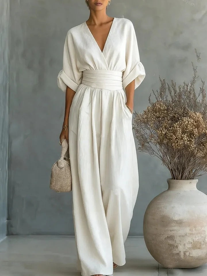 Frontansicht des cremeweißen Jumpsuits mit V-Ausschnitt, breitem Taillenband und weitem Bein, getragen vor grauer Wand neben einer Vase mit Trockenblumen. 1