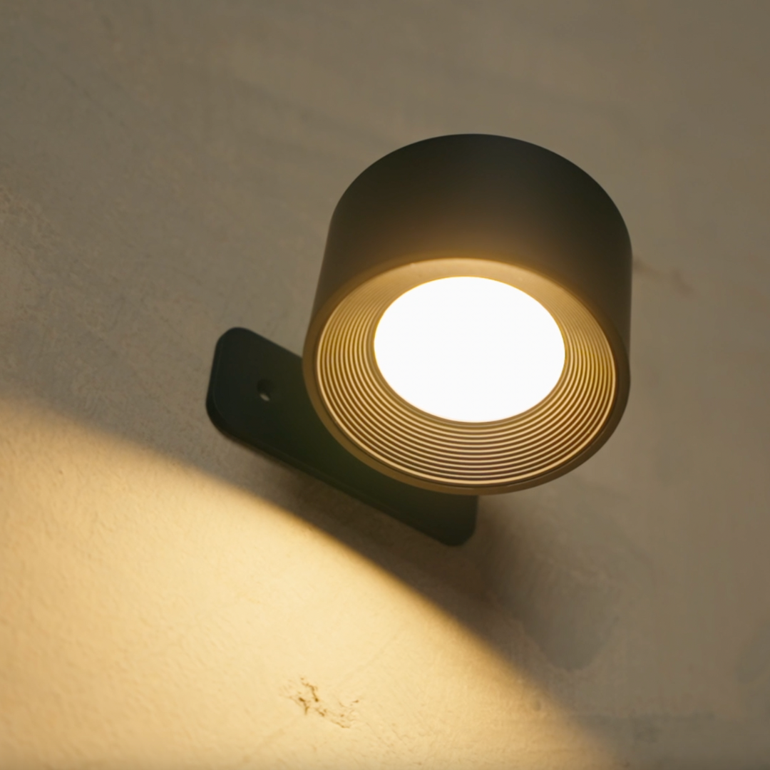 Nahaufnahme einer schwarzen Wandlampe mit hell leuchtender Fläche, gerilltem Innenring und warmem Lichtkegel an der Wand. 16