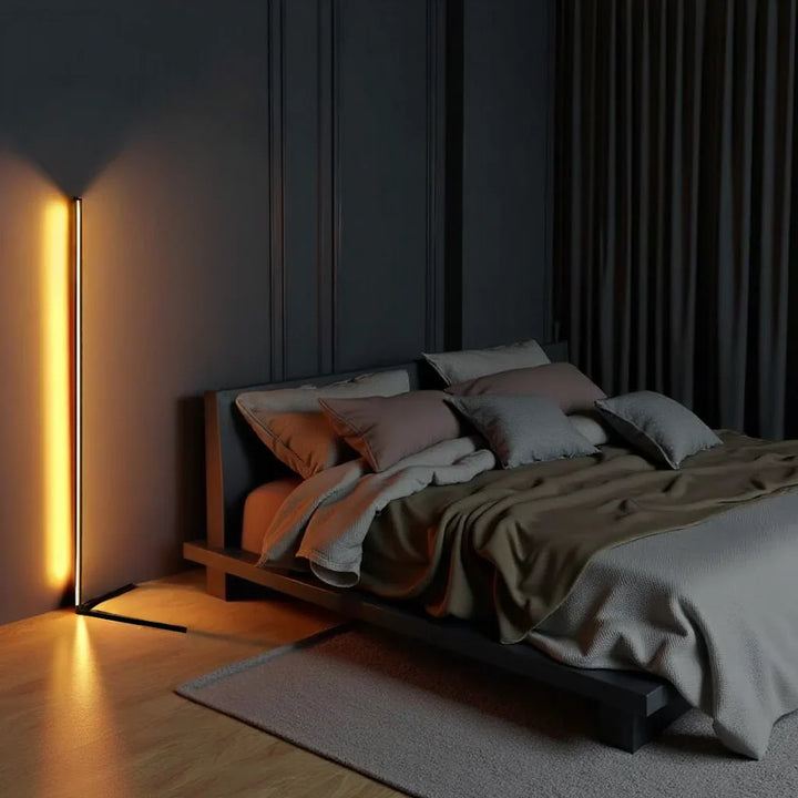 Stehlampe strahlt orange-gelbes Licht an dunkler Wand neben einem niedrigen Bett mit grauen Kissen 2