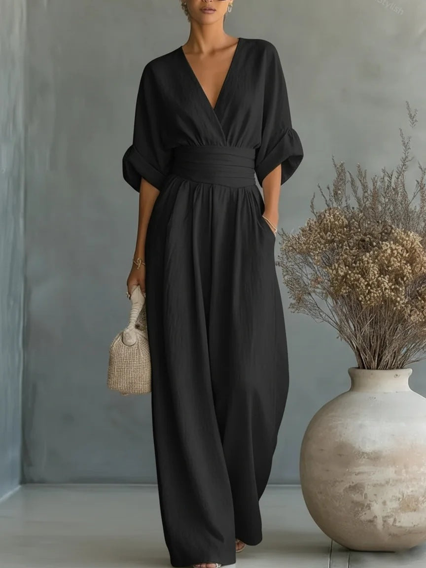 Frontansicht des schwarzen Jumpsuits mit V-Ausschnitt, hoher Taille und umgeschlagenen Ärmeln, getragen mit heller Strohtasche. 5