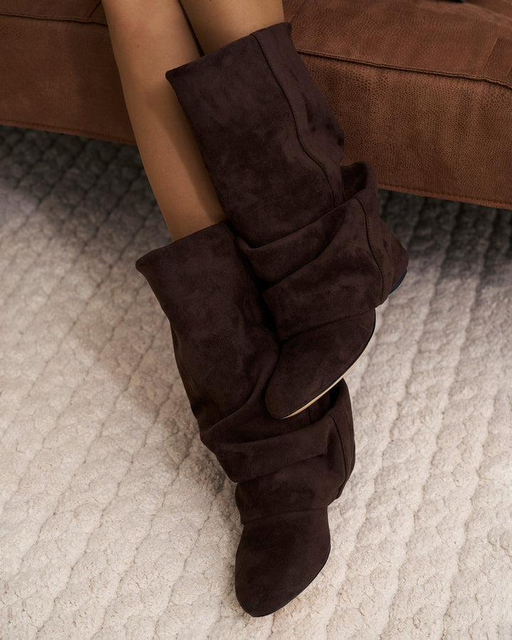 Beine in Strumpfhose sitzen auf einem braunen Sofa und tragen die schokoladenbraunen, gerafften Stiefel; Nahaufnahme auf hellem Teppich. 2