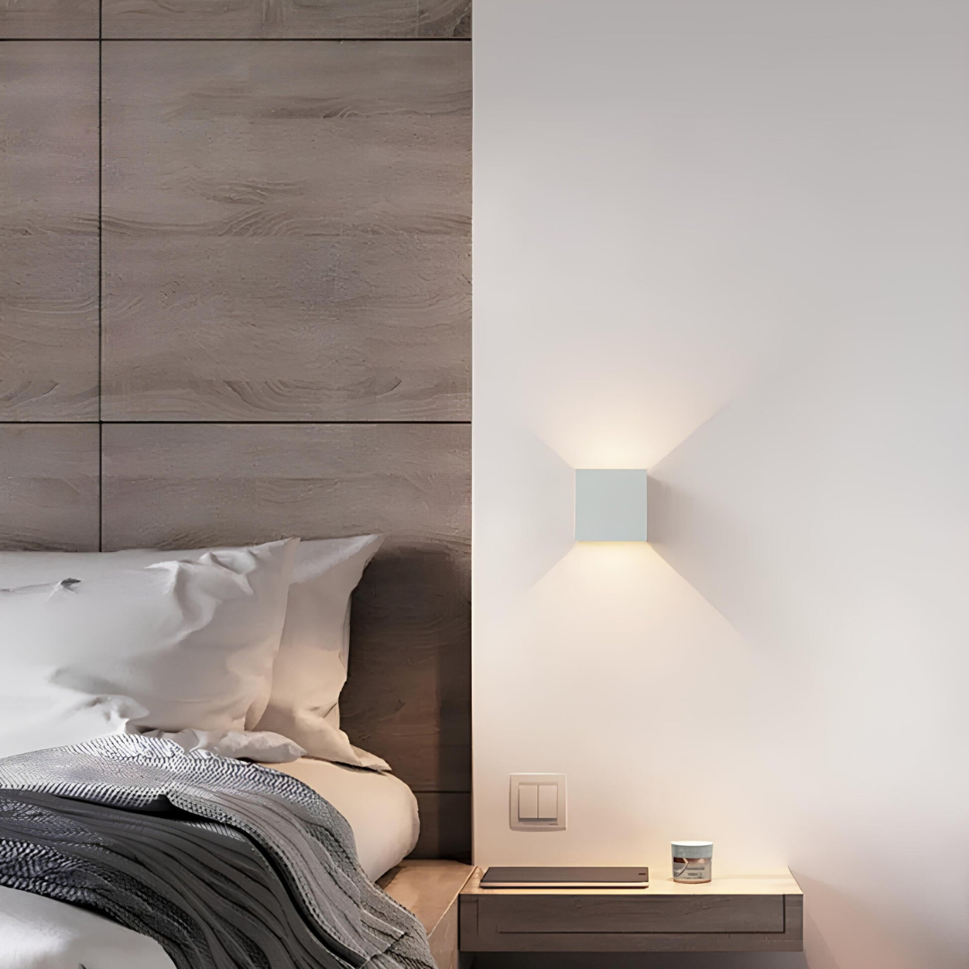 Weiße Würfelleuchte neben einem Bett mit Holzpaneel-Kopfteil, darunter ein Nachttisch mit Tablet und kleiner Dose. 6