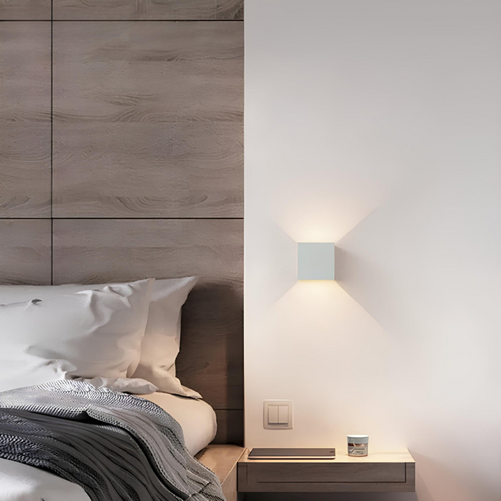 Weiße Würfelleuchte neben einem Bett mit Holzpaneel-Kopfteil, darunter ein Nachttisch mit Tablet und kleiner Dose. 6