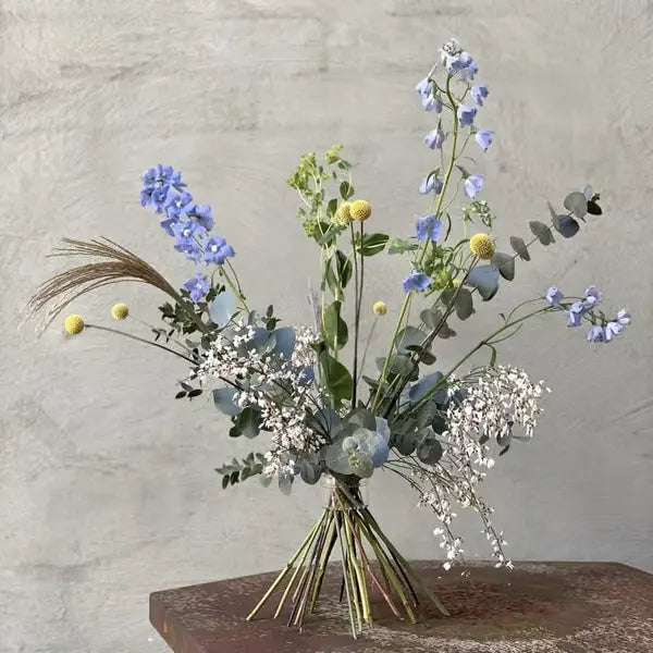 Gebundener Strauß mit blauen Rittersporn-Blüten, gelben Kugelblumen und Eukalyptus, auf einem rostigen Metalltisch vor grauer Wand. 5