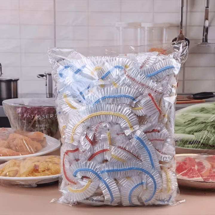 Großer Klarsichtbeutel voller durchsichtiger Abdeckungen mit blauen, gelben und roten Gummibändern vor Speisen in einer Küche. 1