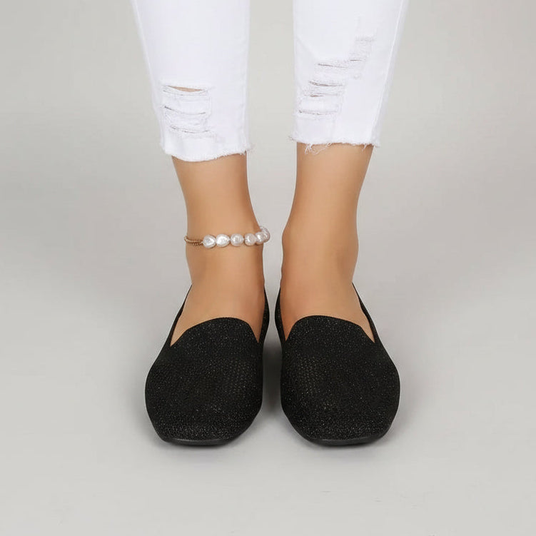 Frontansicht schwarzer glitzernder Slip-ons an den Füßen einer Frau mit weißer Jeans und Perlen-Fußkettchen vor grauem Hintergrund. 2