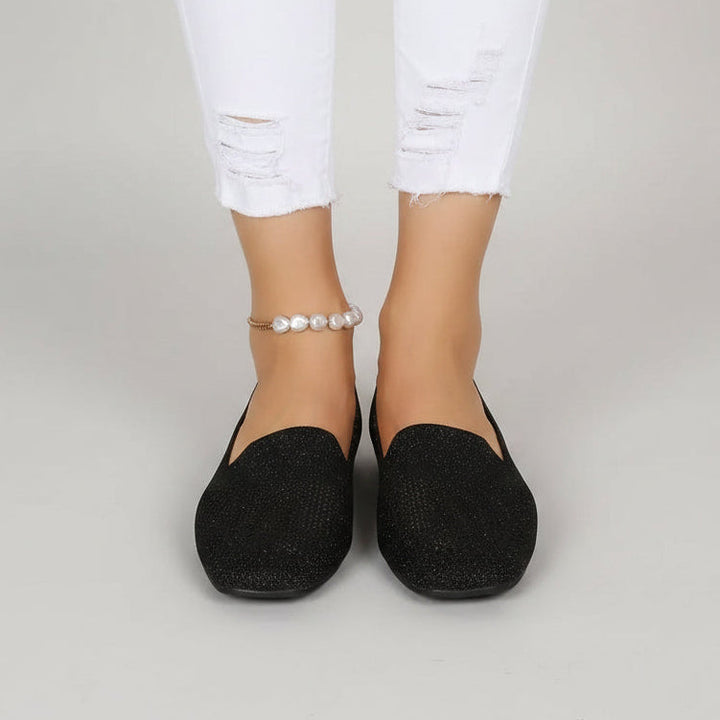 Frontansicht schwarzer glitzernder Slip-ons an den Füßen einer Frau mit weißer Jeans und Perlen-Fußkettchen vor grauem Hintergrund. 2