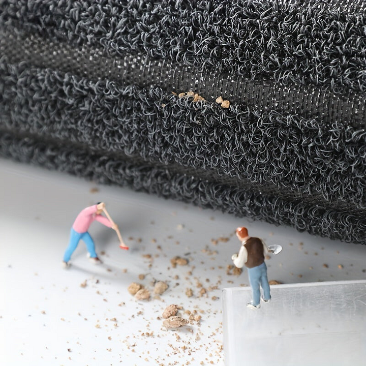 Aufgerollte Mattenkante mit Blick auf Faserschlingen und Gewebeträger, davor Miniaturfiguren, die herabgefallenen Schmutz wegkehren. 6